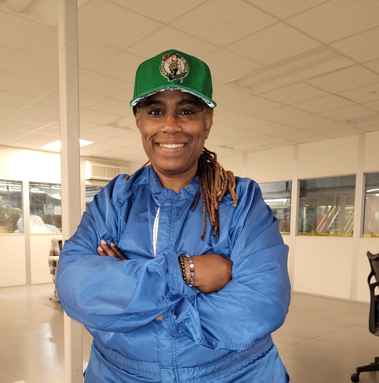GM employee wearing a green hat and a blue jumpsuit stands smiling inside a conference room with arms crossed over her chest.