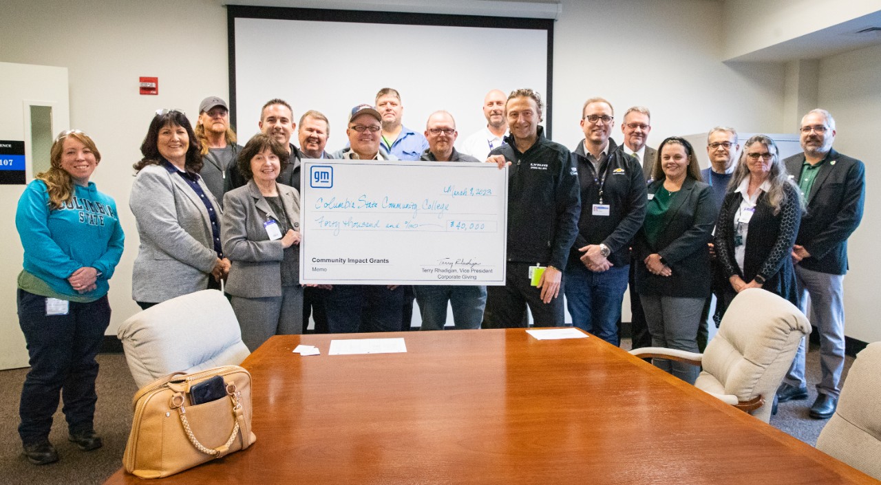 Group of GM & UAW employees & Columbia State college officials stand in front of a brown desk holding a giant white GM check
