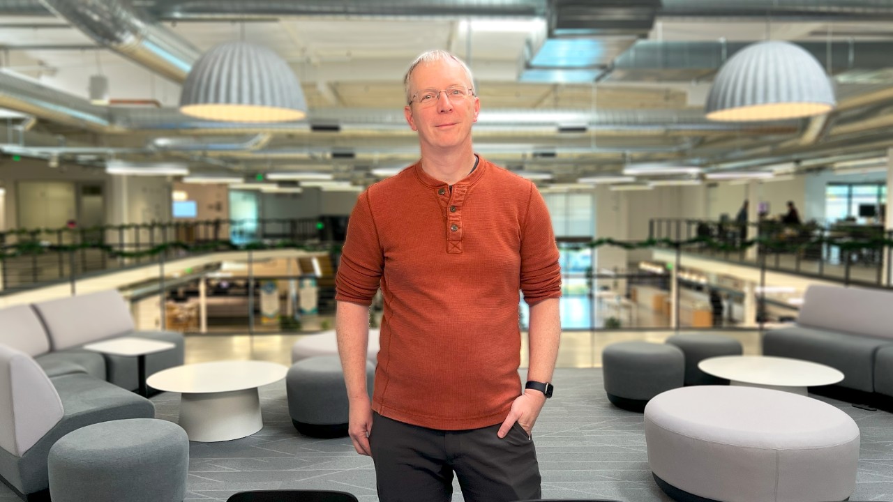 Fraser Graham smiles while standing center-frame in a modern office setting wearing an orange shirt and dark pants.