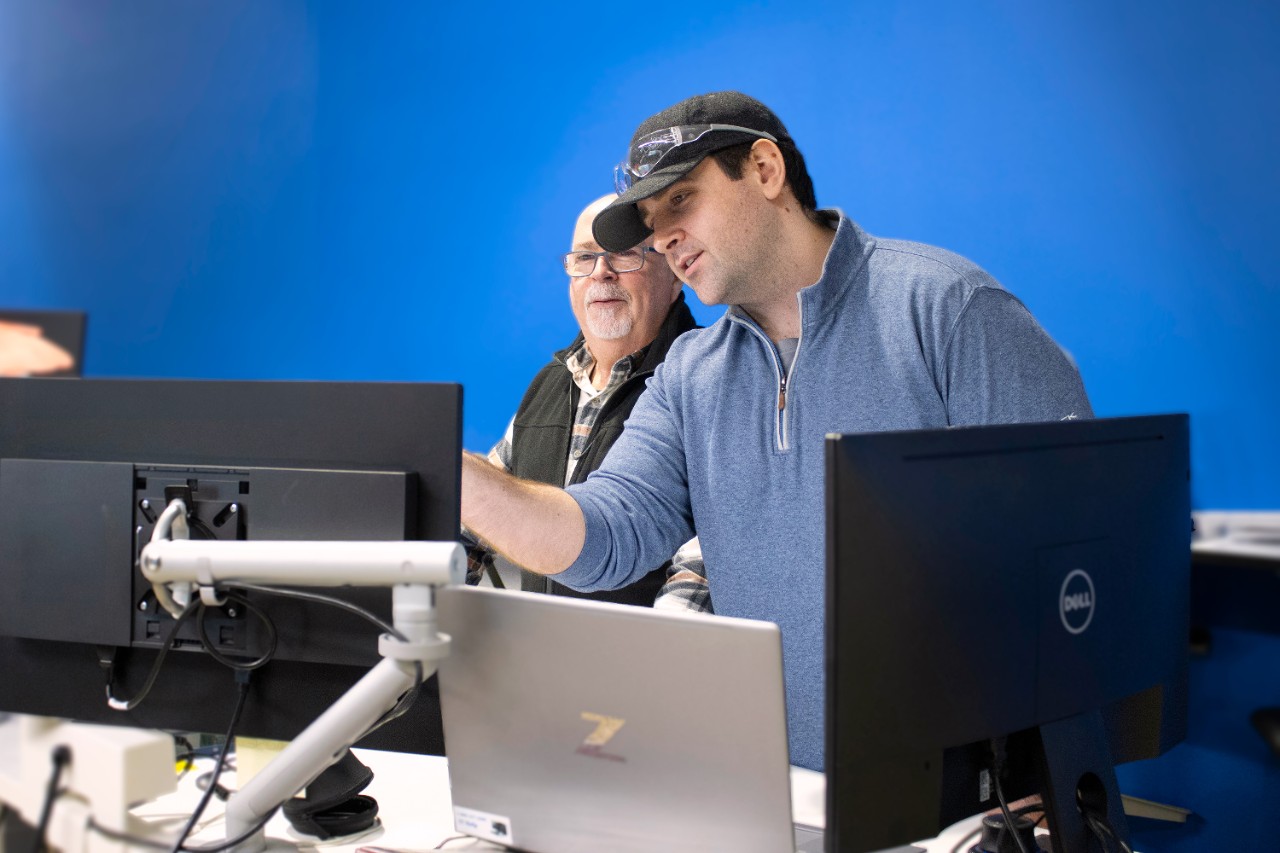 Max Sikorski wears a blue quarter-zip, jeans and a black baseball cap standing beside VR Specialist Kevin LaPrise behind