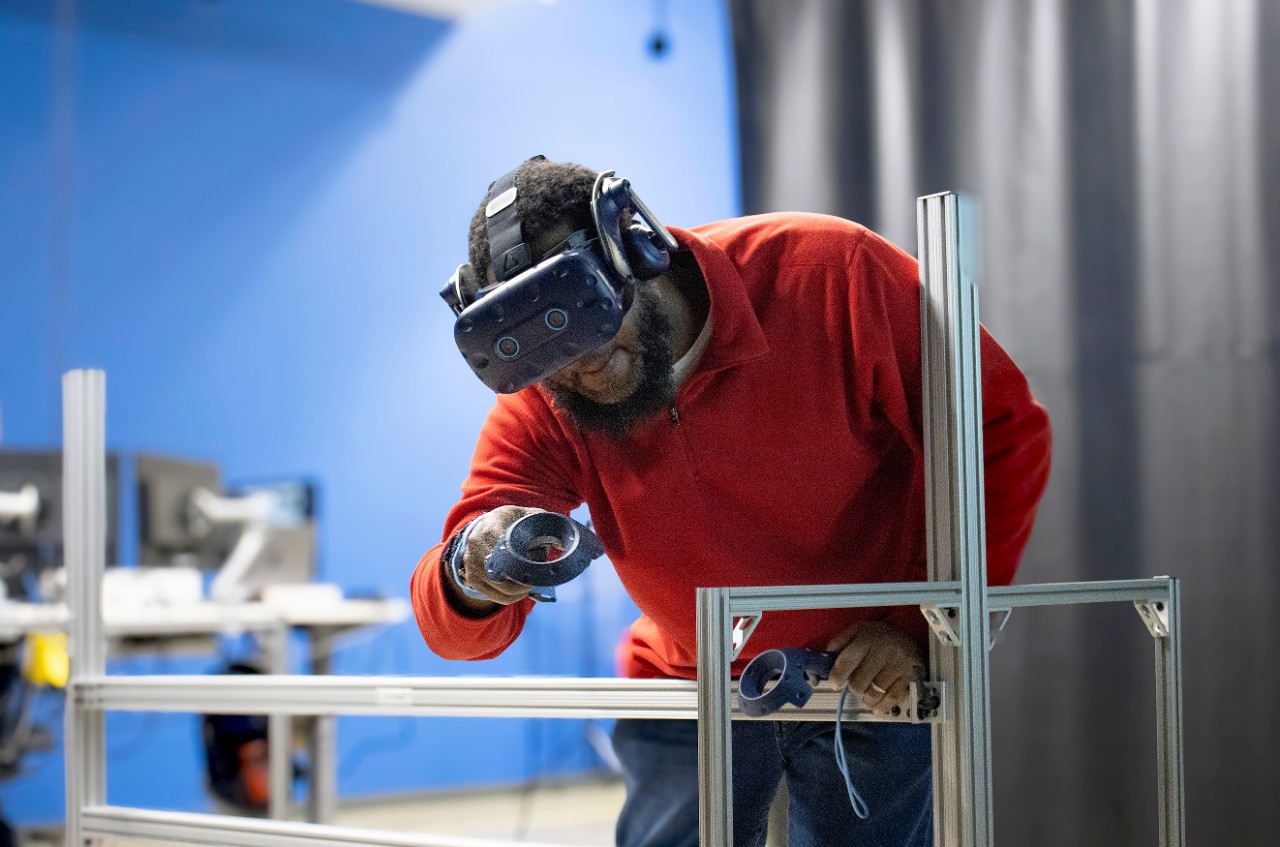 VR Operator Dwaine Carswell wears a red quarter-zip and jeans while using a virtual reality headset and holding two VR wands