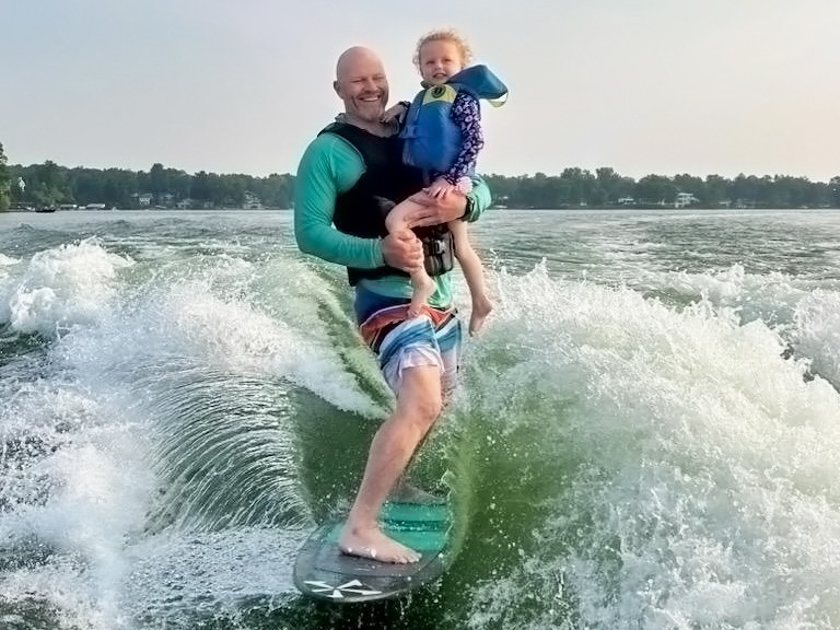 Man holding a child while wake surfing on a lake.