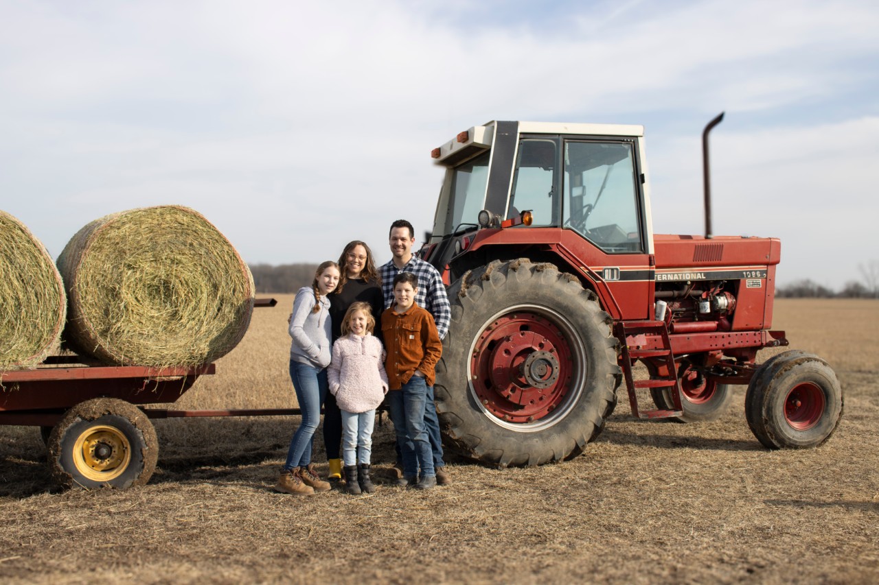 IMG_4702 Hillary Lange, her husband Rob, and their three children pose in front of a red tractor and trailer full of hay bales.