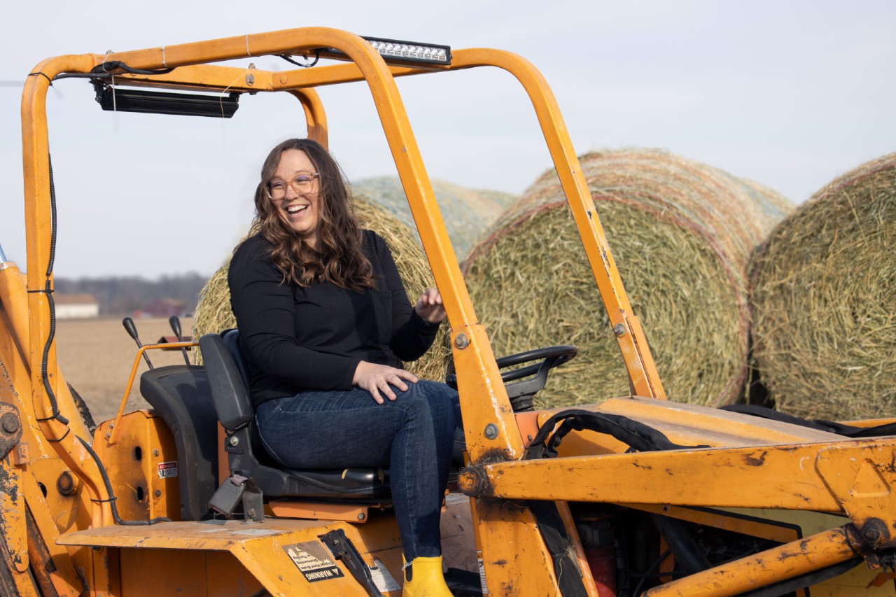 Hillary Lange wears a black shirt and jeans while driving a yellow tractor in front of hay bales.