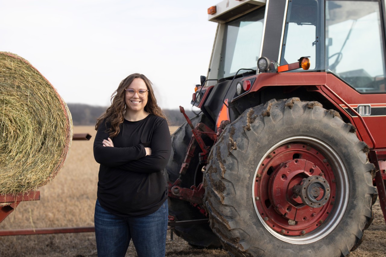 Hillary Lange wears a black shirt and jeans while posing in front of a red tractor and trailer full of hay bales. 