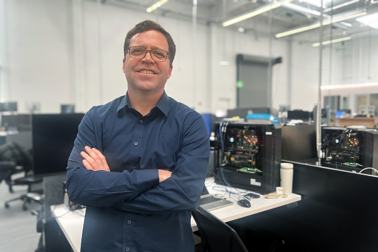 A man in a blue shirt standing in an office with his arms crossed.