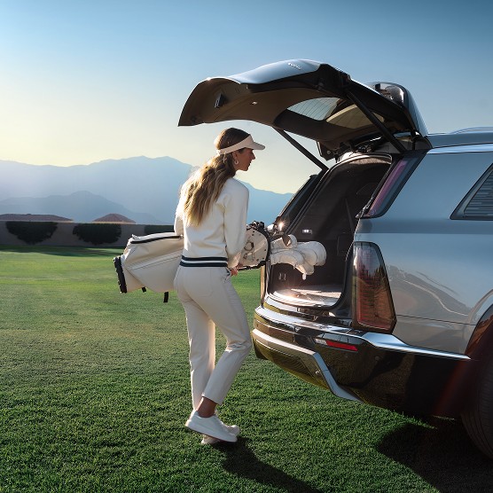 Woman loading golf clubs into the rear cargo area of a Cadillac VISTIQ luxury SUV parked near a golf course at sunrise.