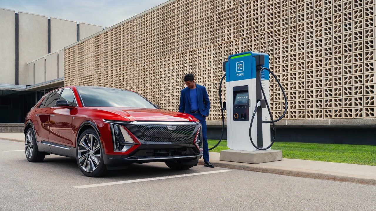 A man in a blue suit standing by a red Cadillac LYRIQ electric SUV parked at a GM EVgo charging station.