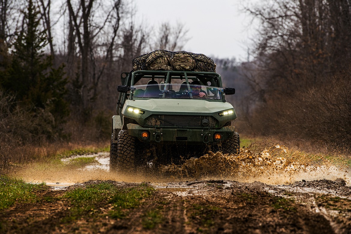 Front of a man operating a military-grade, open-frame vehicle in matte army green with rugged tires on dirt road.